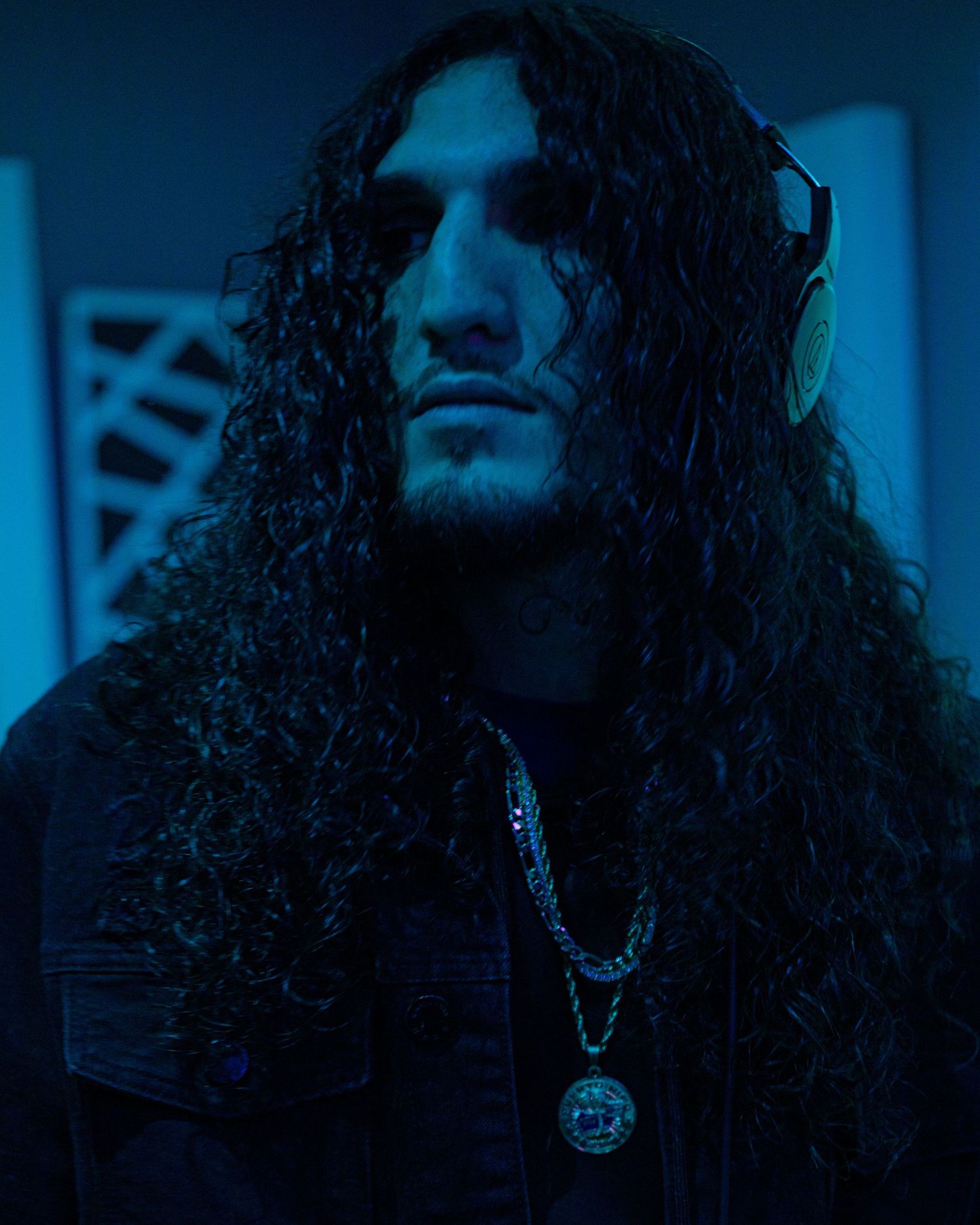 Musician with long curly hair wearing headphones and chain necklace in blue-lit studio.