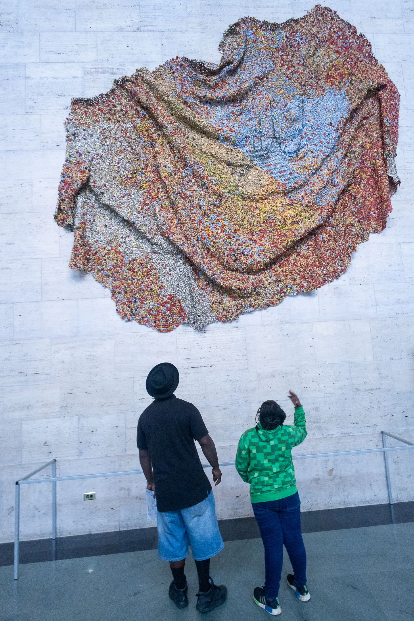Two museum visitors admire a large textile wall sculpture made of colorful recycled materials.