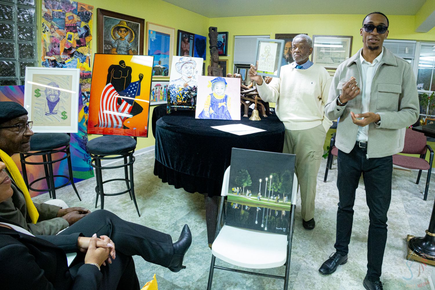 Two men present colorful artwork and paintings at an indoor art gallery exhibition event.