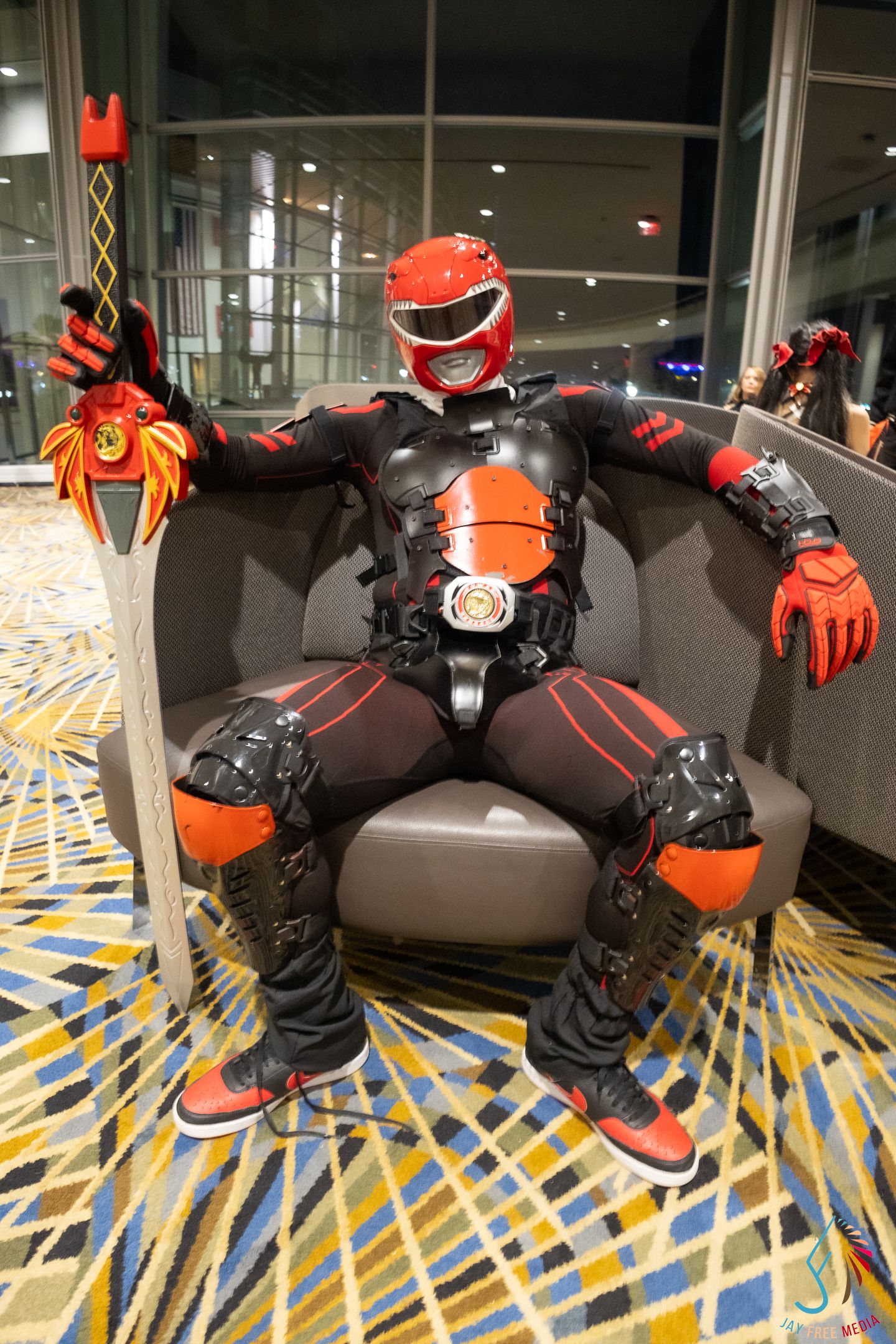 Person in red and black Power Rangers cosplay costume seated in chair at comic convention.