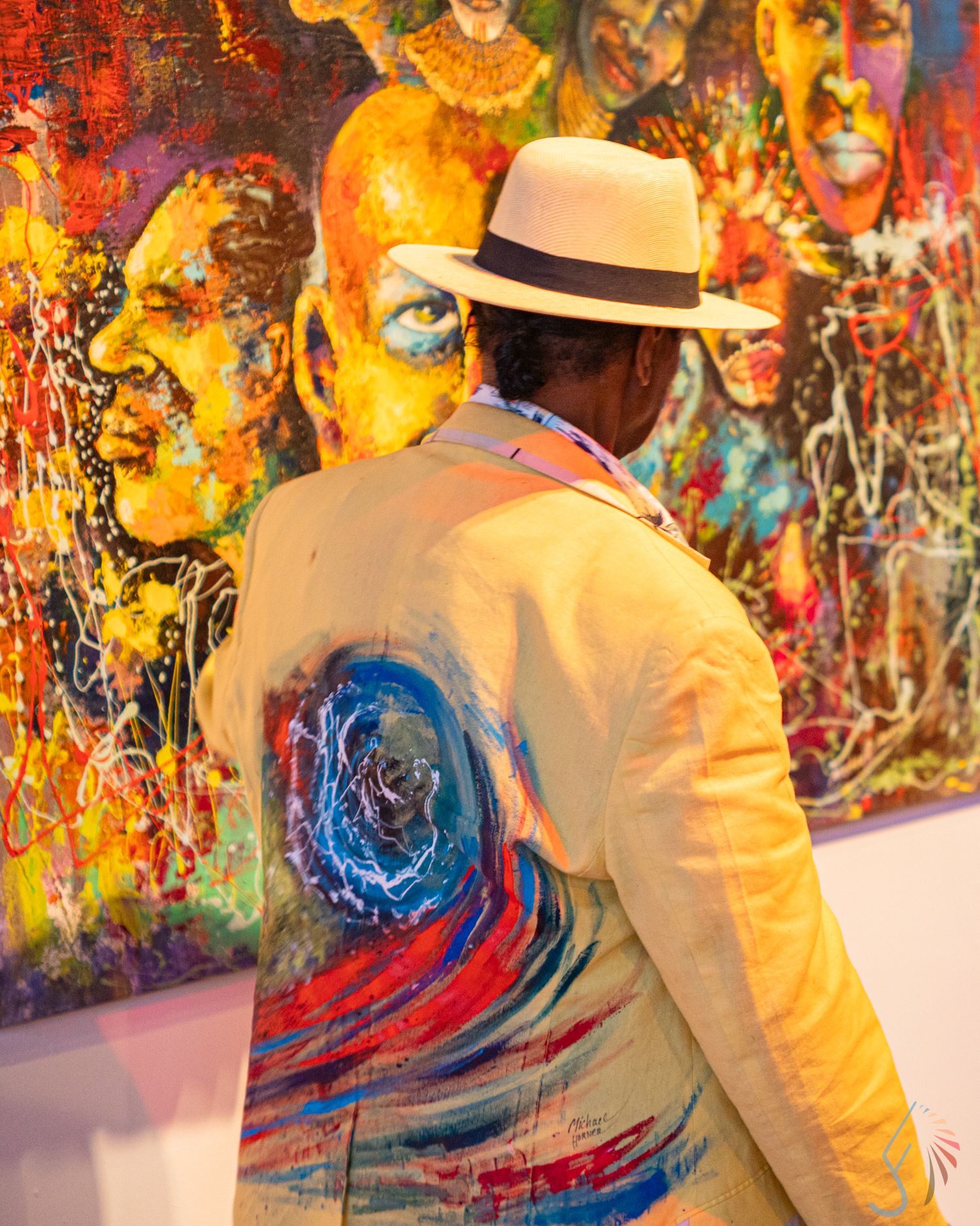 Man in white hat and painted jacket views vibrant colorful portrait artwork at an art gallery exhibition.