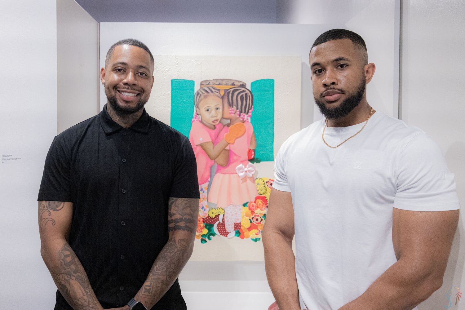 Two men pose together at an art gallery with colorful paintings displayed on the white walls behind them.