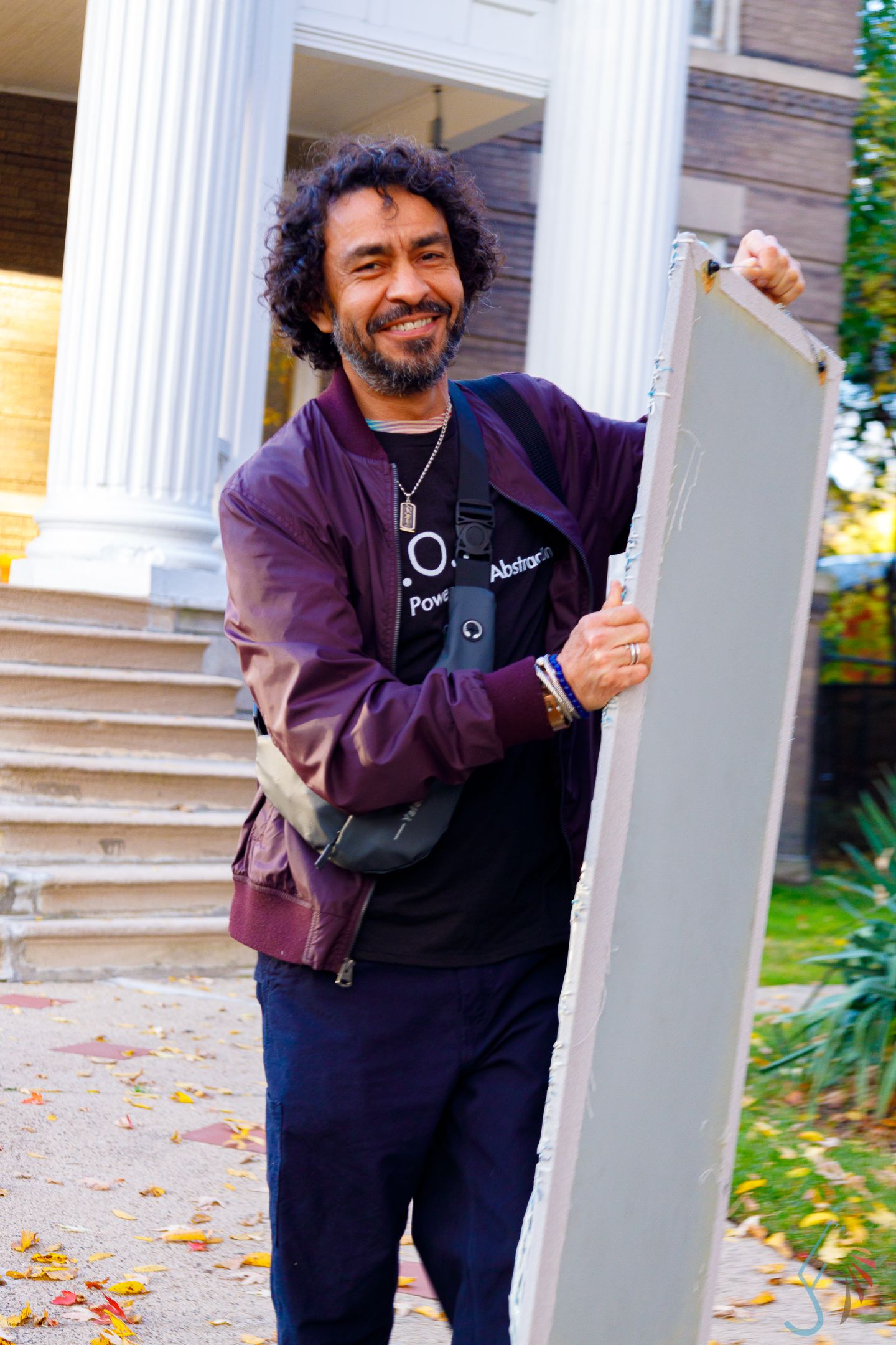 Smiling man with curly hair holding a canvas board outdoors near white columns in autumn.