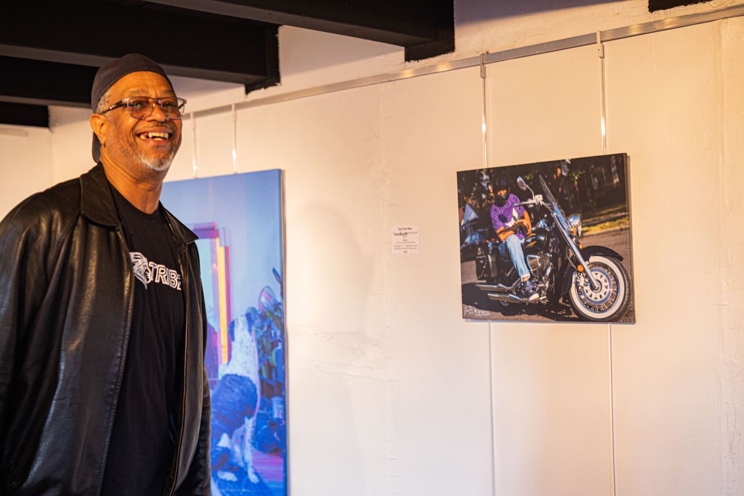 Man in black leather jacket smiling beside motorcycle photography prints at an art gallery exhibition.