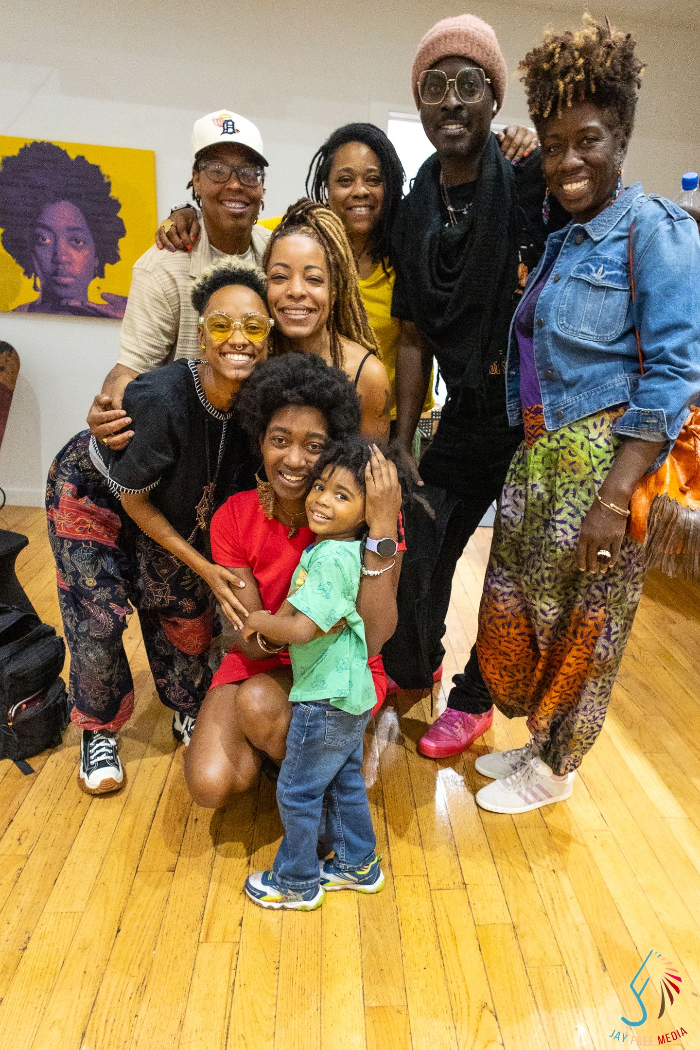 A joyful group of Black adults and a child posing together indoors at a cultural event, with colorful artwork on the wall behind them.