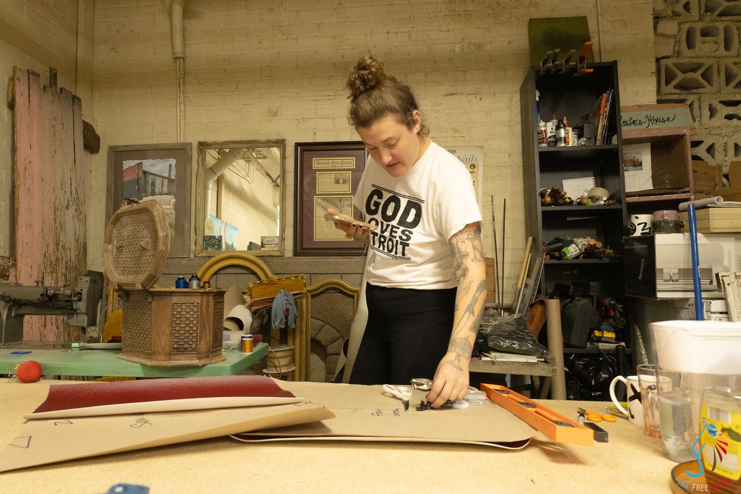 Woman in God t-shirt working with materials at a cluttered workshop table in an art studio.