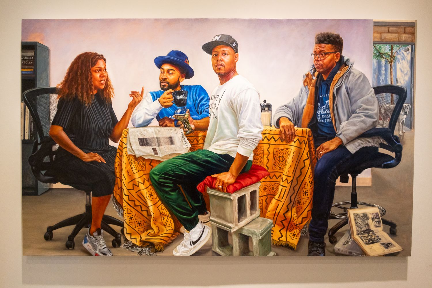 Colorful painting of four people sitting together in an office setting, displayed on a white gallery wall.