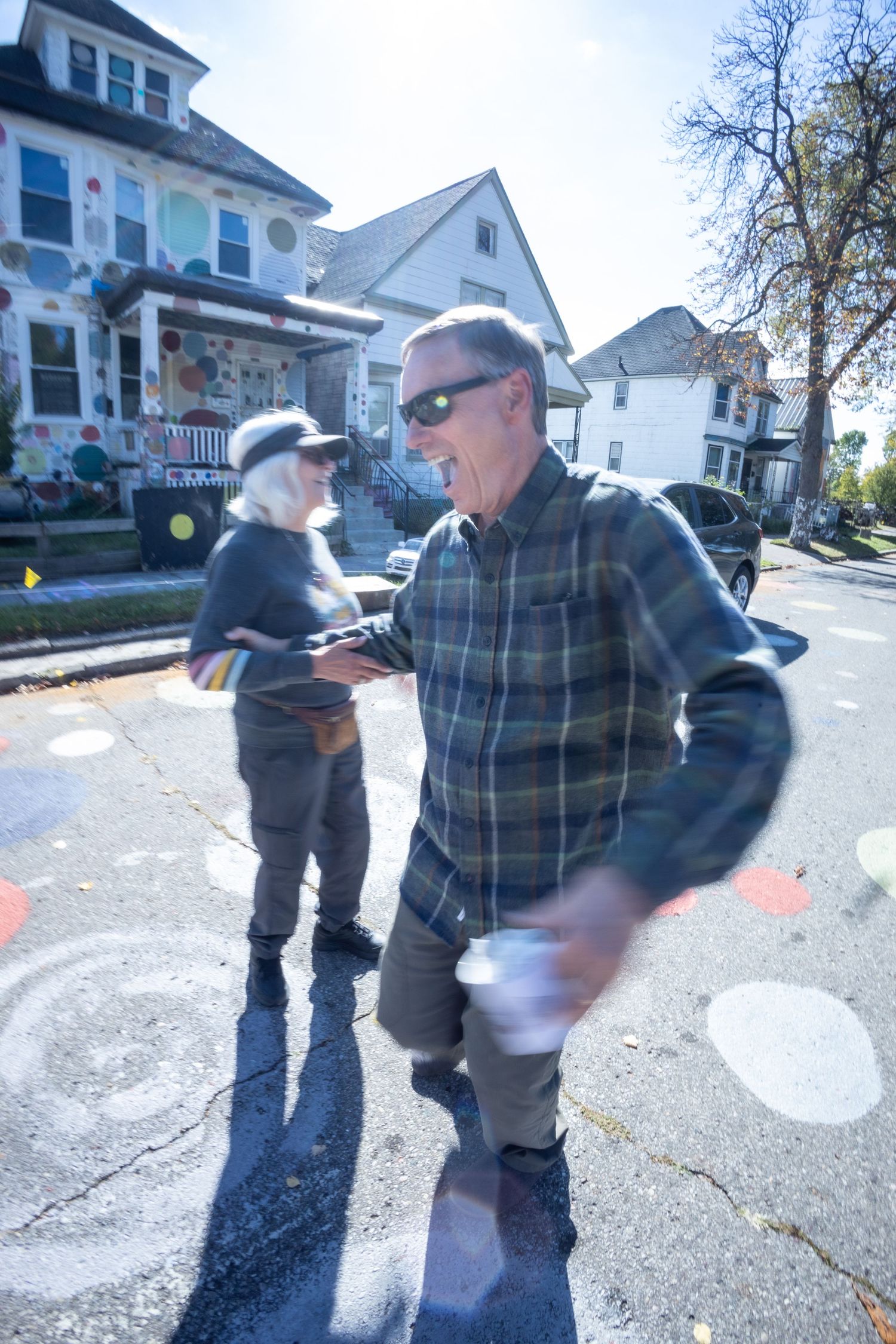 Two people walk and smile on a colorful street with polka-dot painted houses in the background.