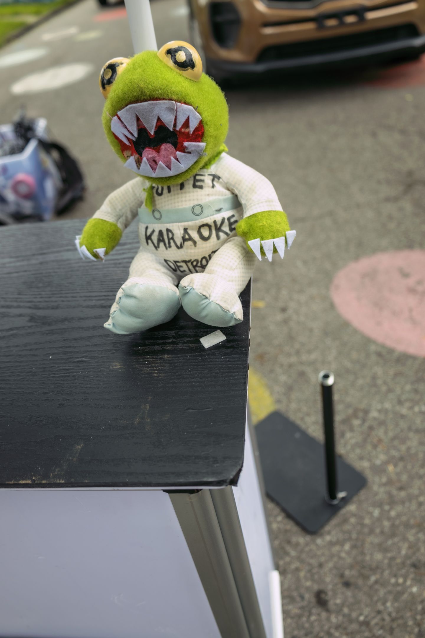 Green frog puppet with sharp teeth wearing karaoke Detroit outfit sits on black table outdoors.