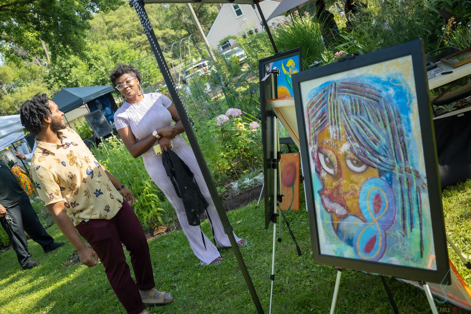 Two people admire a colorful portrait painting on an easel at an outdoor art event in a lush garden setting.