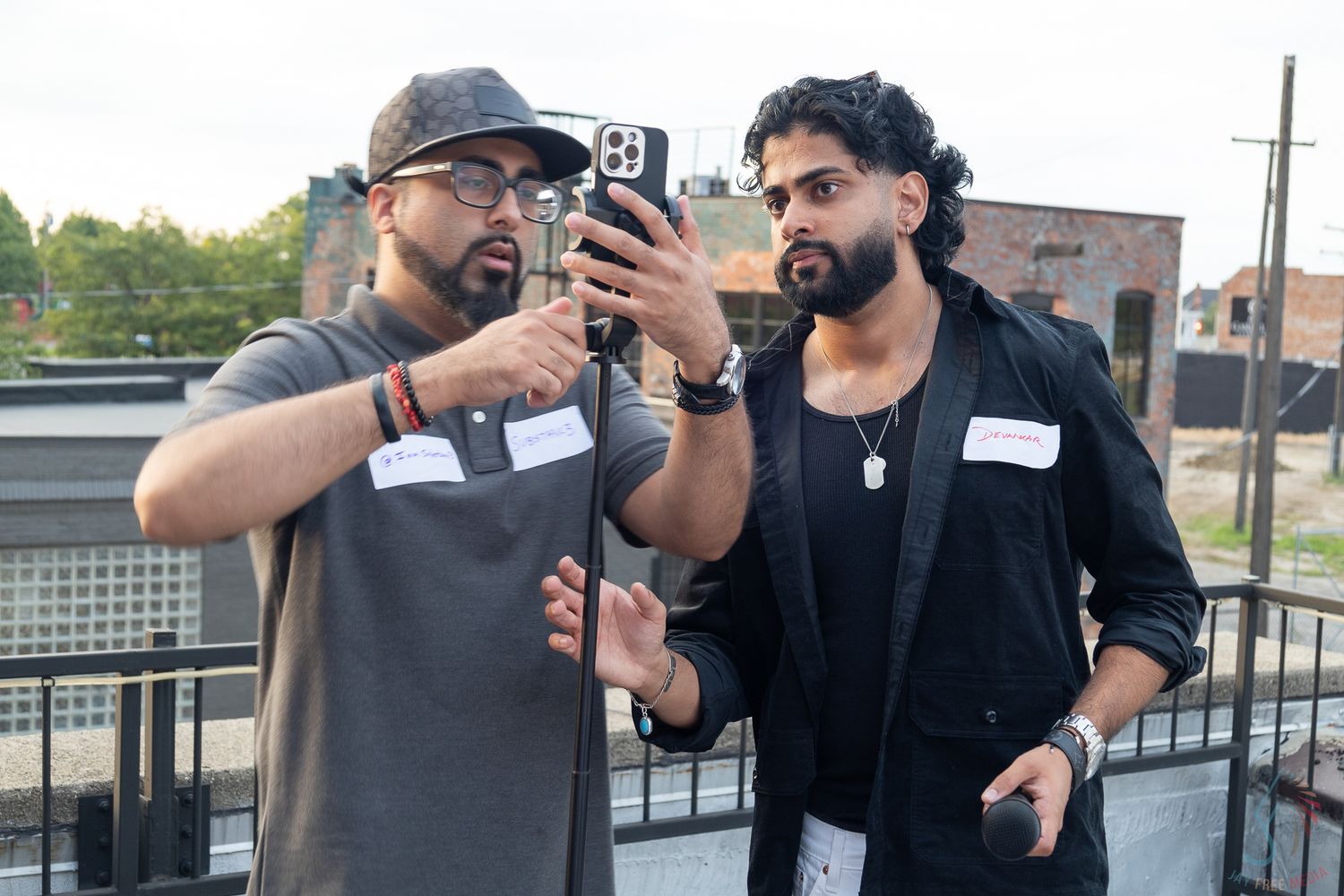 Two men review footage on smartphone at outdoor rooftop networking event.