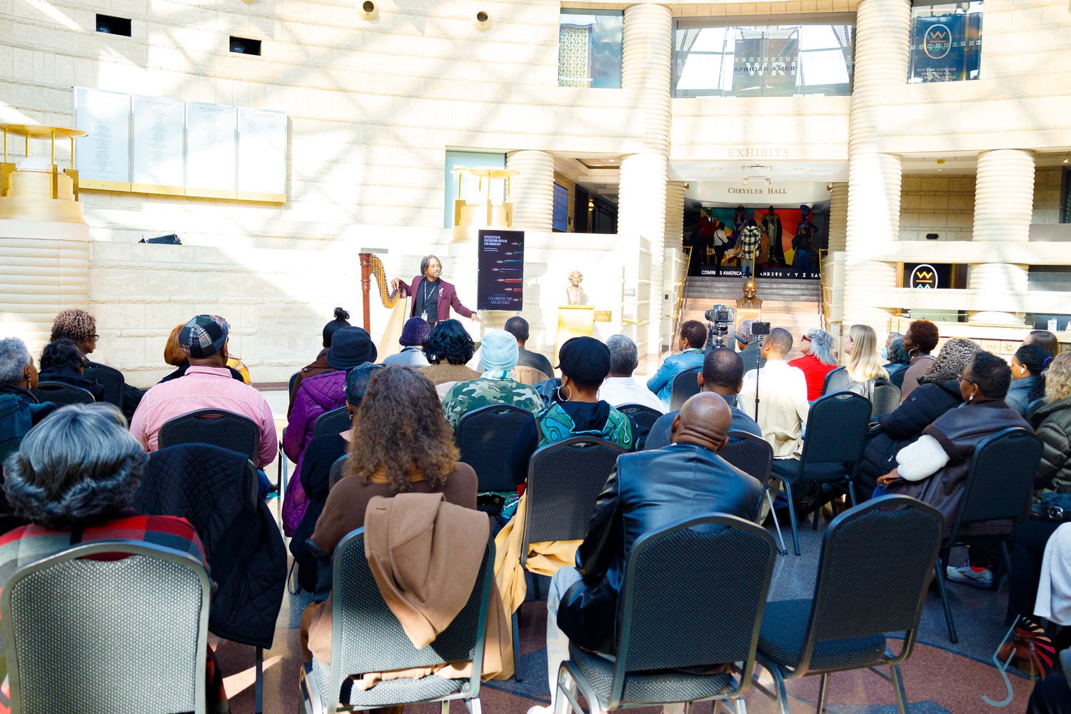 Audience seated in chairs watching a harpist perform in a sunlit atrium during a live indoor cultural event.