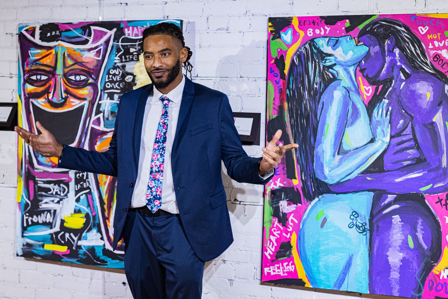 Man in blue suit gestures beside vibrant neo-expressionist paintings at an art gallery exhibition.