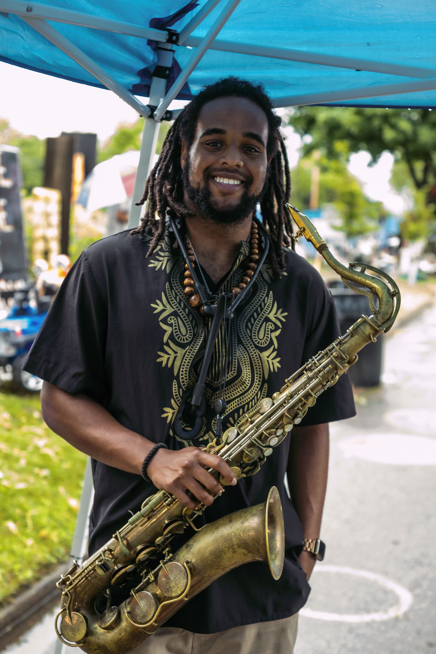 Smiling man with dreadlocks holding a brass saxophone outdoors at a street festival under a blue tent.