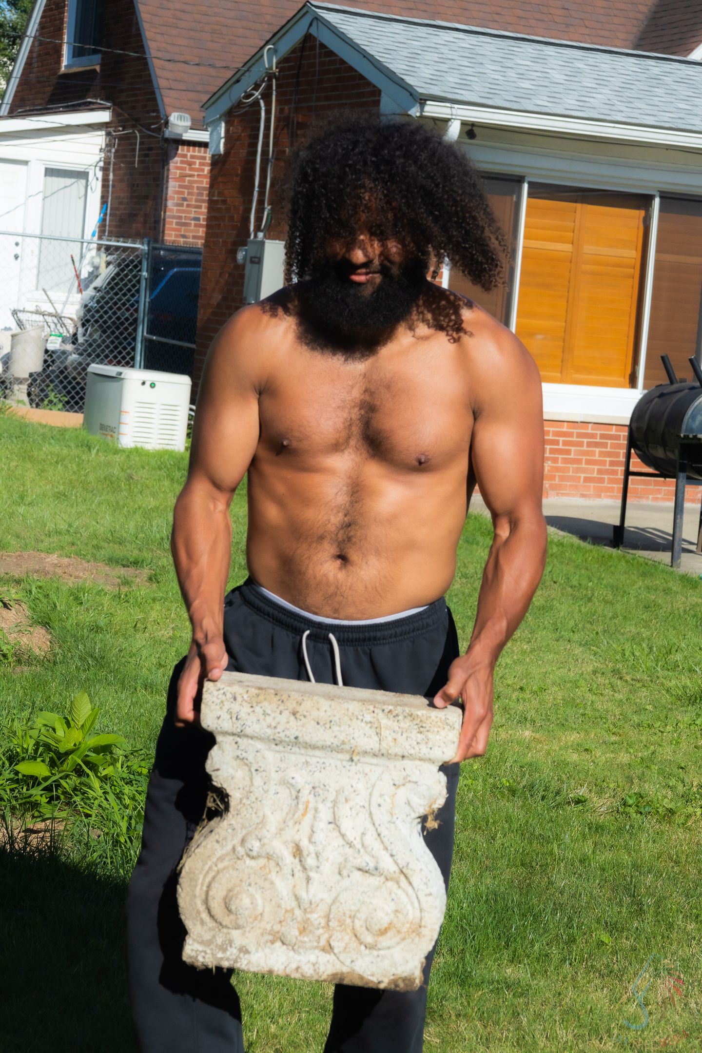 Muscular shirtless man with curly hair and beard holding a decorative stone architectural piece outdoors.