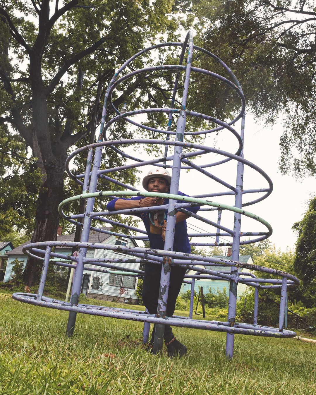 Child playing on a spiral metal playground structure in a green park with large trees in the background.