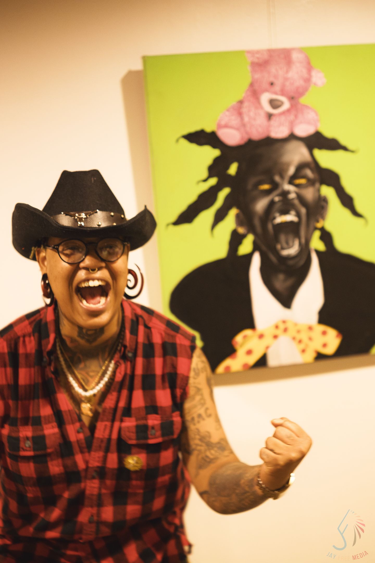 An expressive woman in a cowboy hat and plaid shirt poses in front of a vibrant pop art painting.