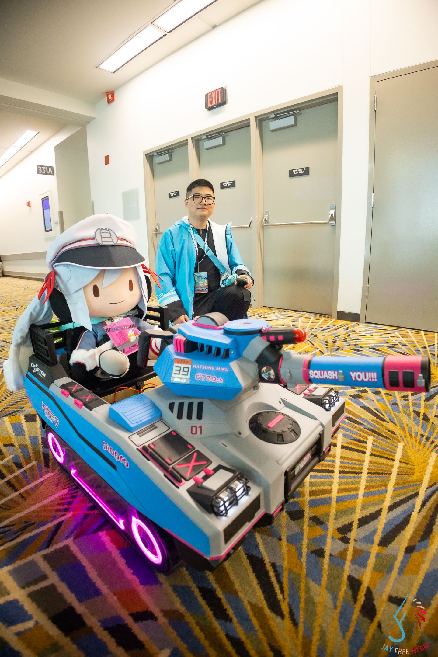 Cosplayer in teal outfit poses with detailed D.Va mech prop and plush doll at anime convention.