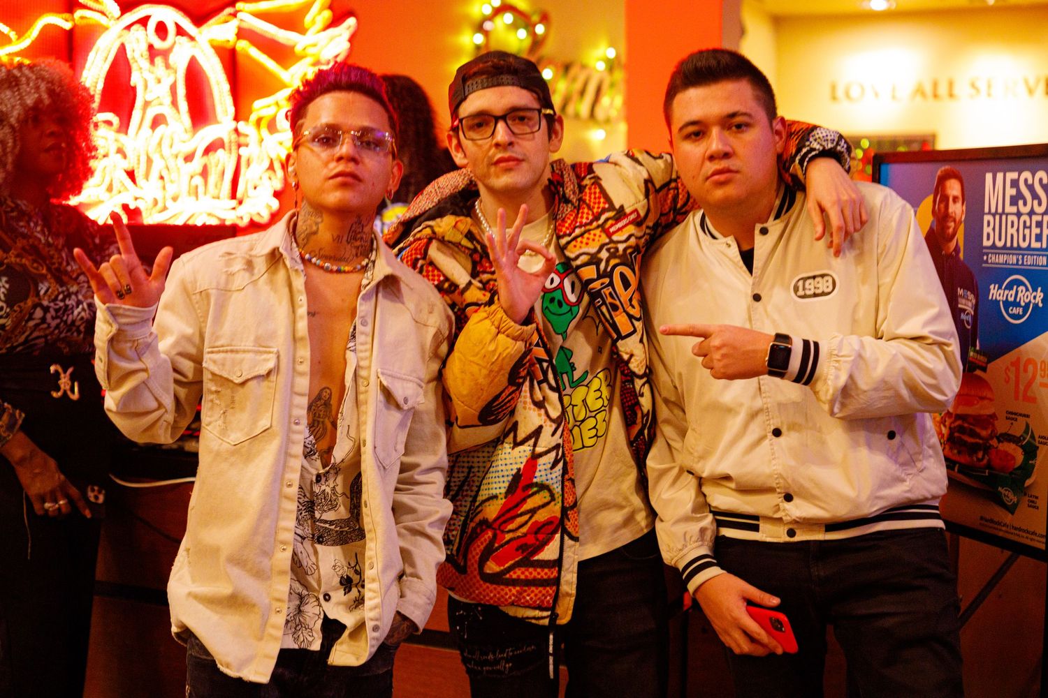 Three young men pose together in front of a Hard Rock Cafe neon sign at a lively night event.