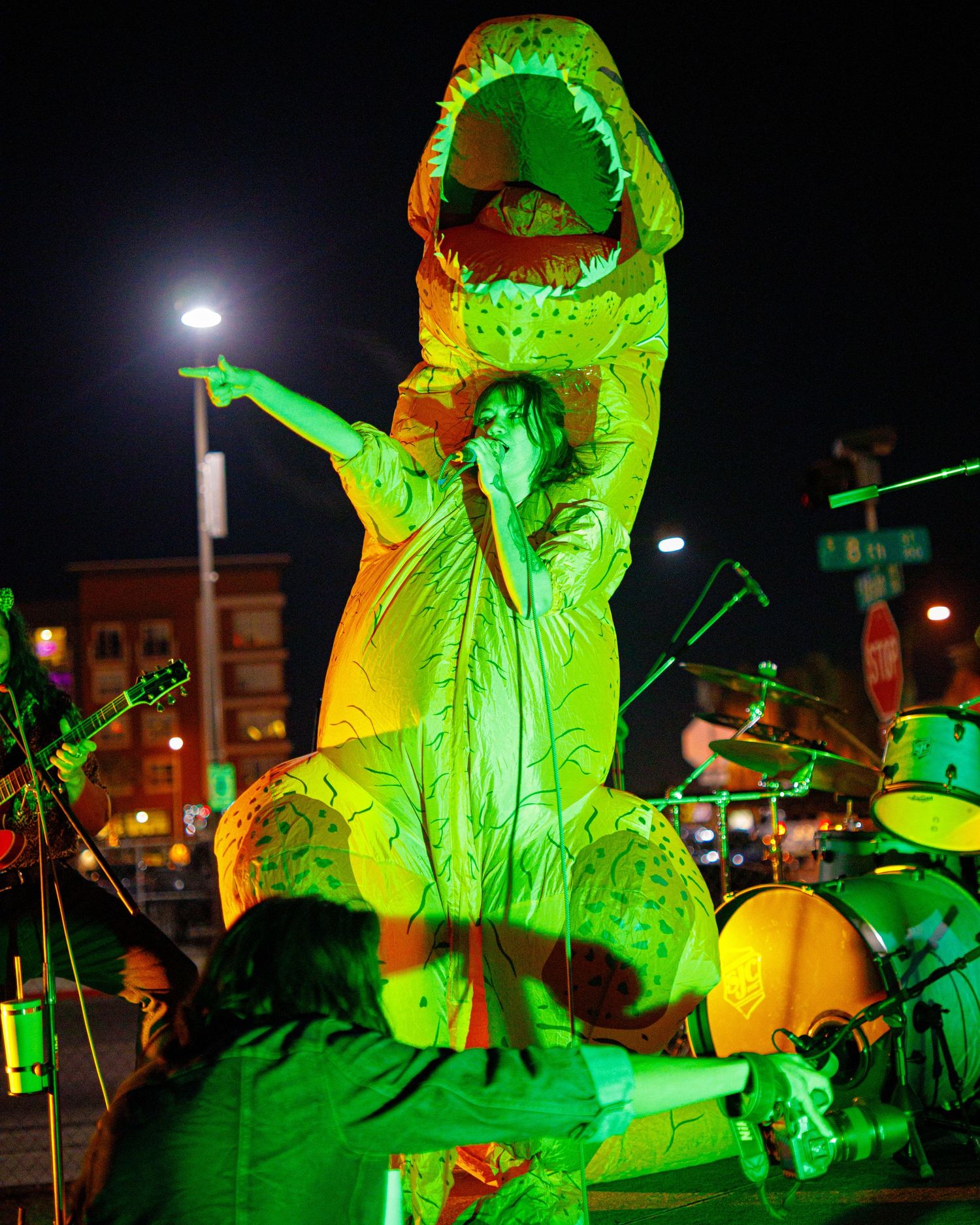 Performer in green T-Rex dinosaur costume singing on stage under green lights at night concert.