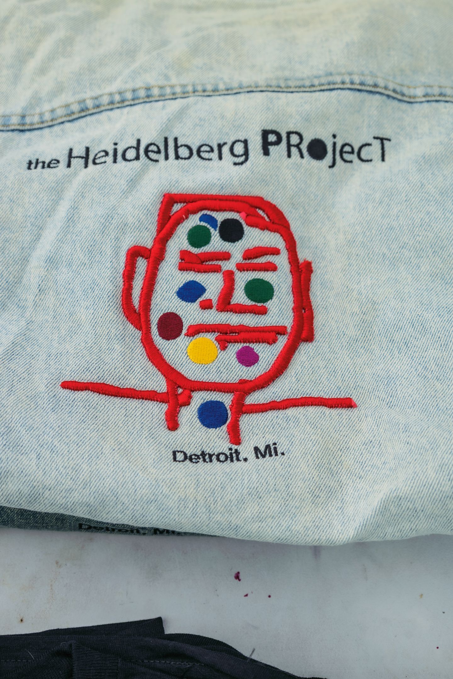 Denim jacket with embroidered Heidelberg Project logo featuring colorful face design, Detroit, MI.