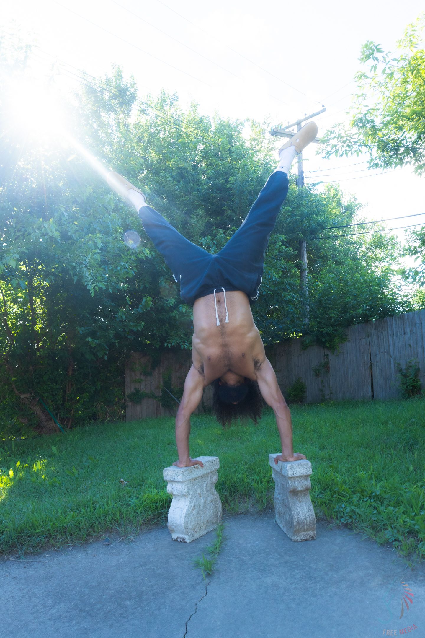 Shirtless man performing a handstand on two stone pedestals outdoors in a backyard with green trees.