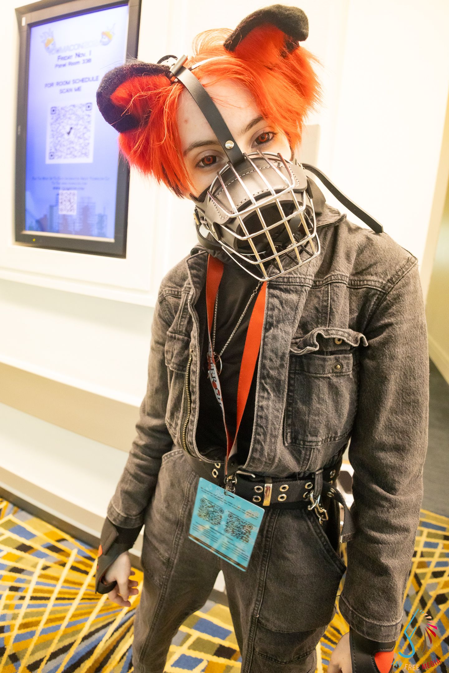 Cosplayer with orange hair wearing a metal muzzle mask and denim jumpsuit at a convention.