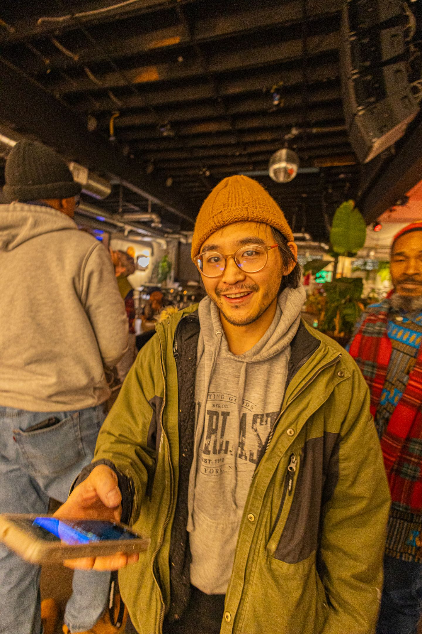 Smiling young man wearing glasses, brown beanie, and olive jacket holds a phone inside a busy venue.