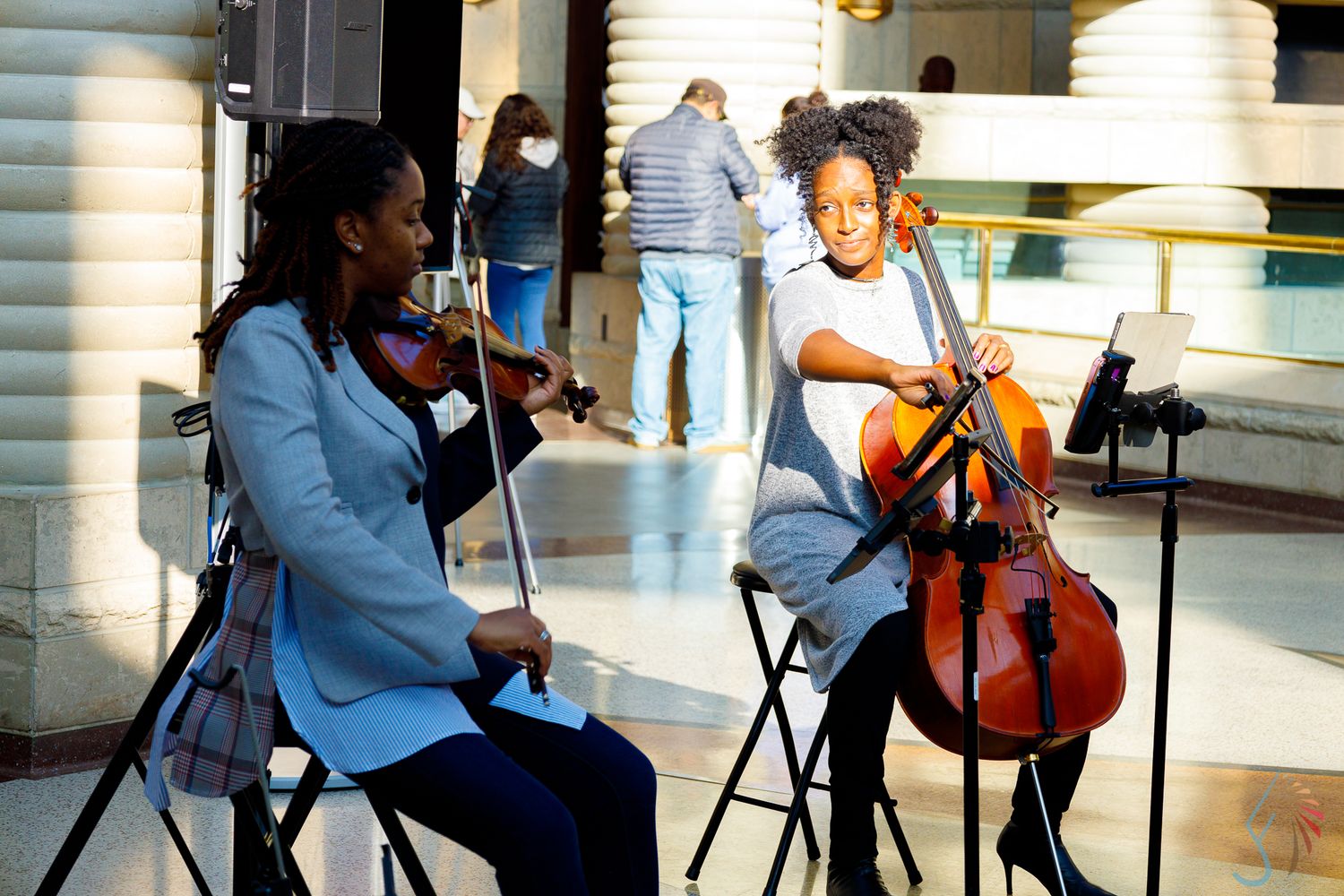 Two young women playing violin and cello in a bright indoor venue, wearing light blue blazers.