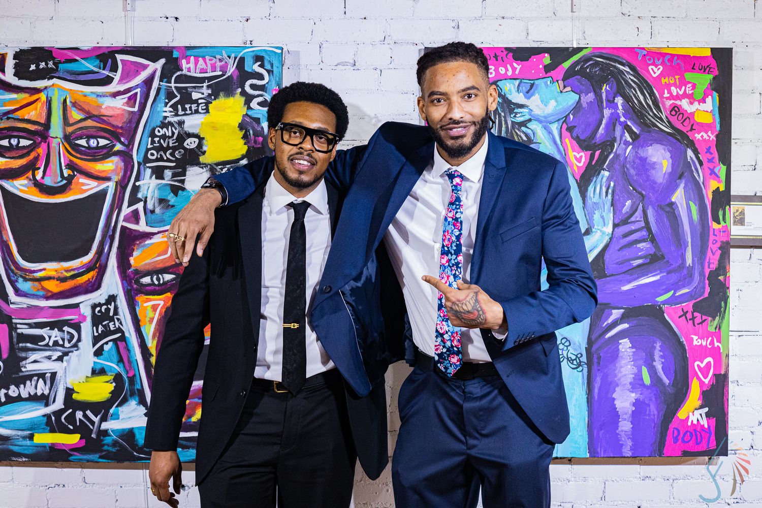Two stylish men in suits pose in front of vibrant colorful graffiti art paintings.
