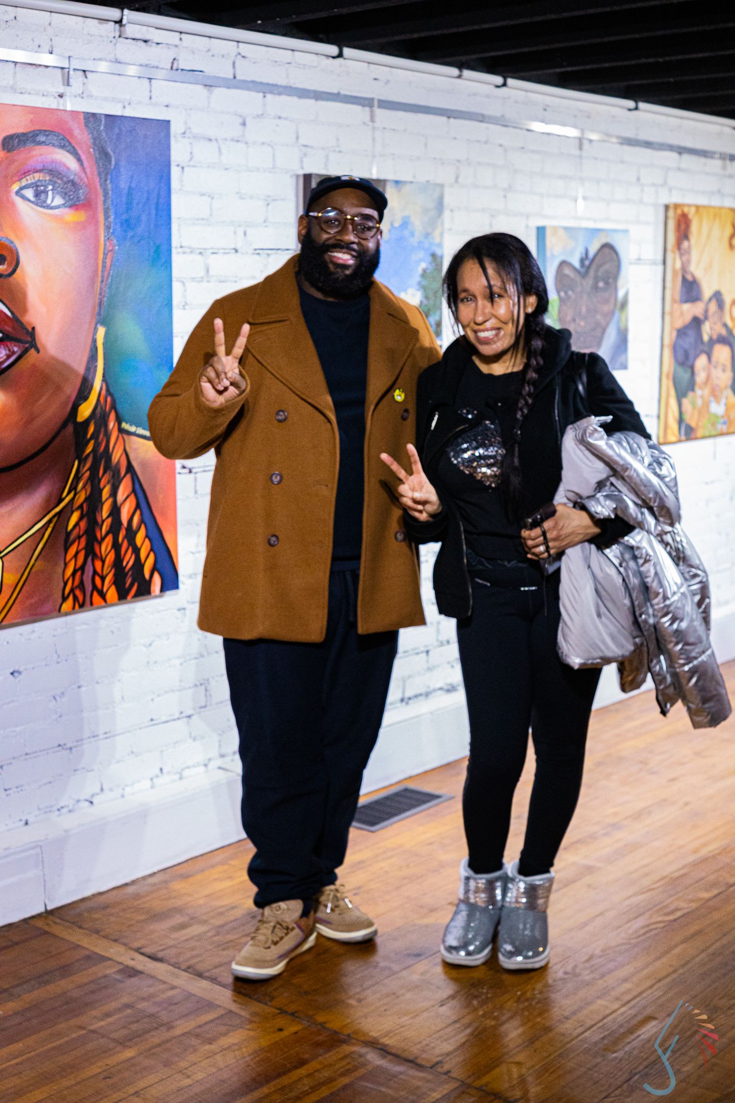 Two people posing with peace signs at an art gallery with colorful portrait paintings on white brick walls.