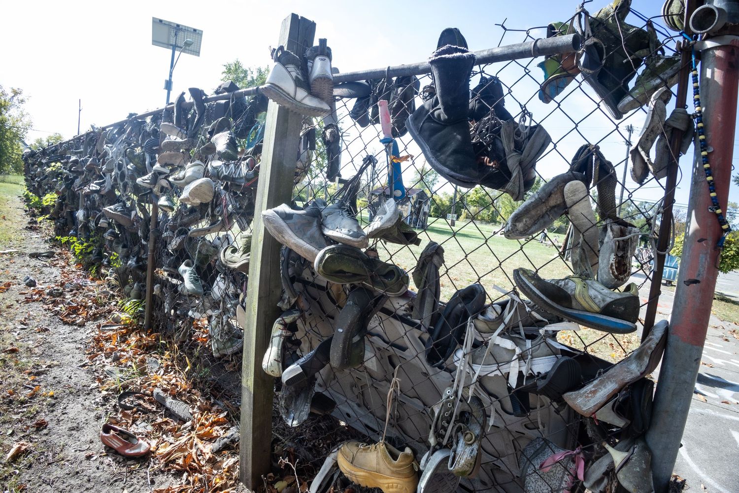 Shoes and boots covering a chain-link fence along a grassy roadside, creating an unusual outdoor art installation.