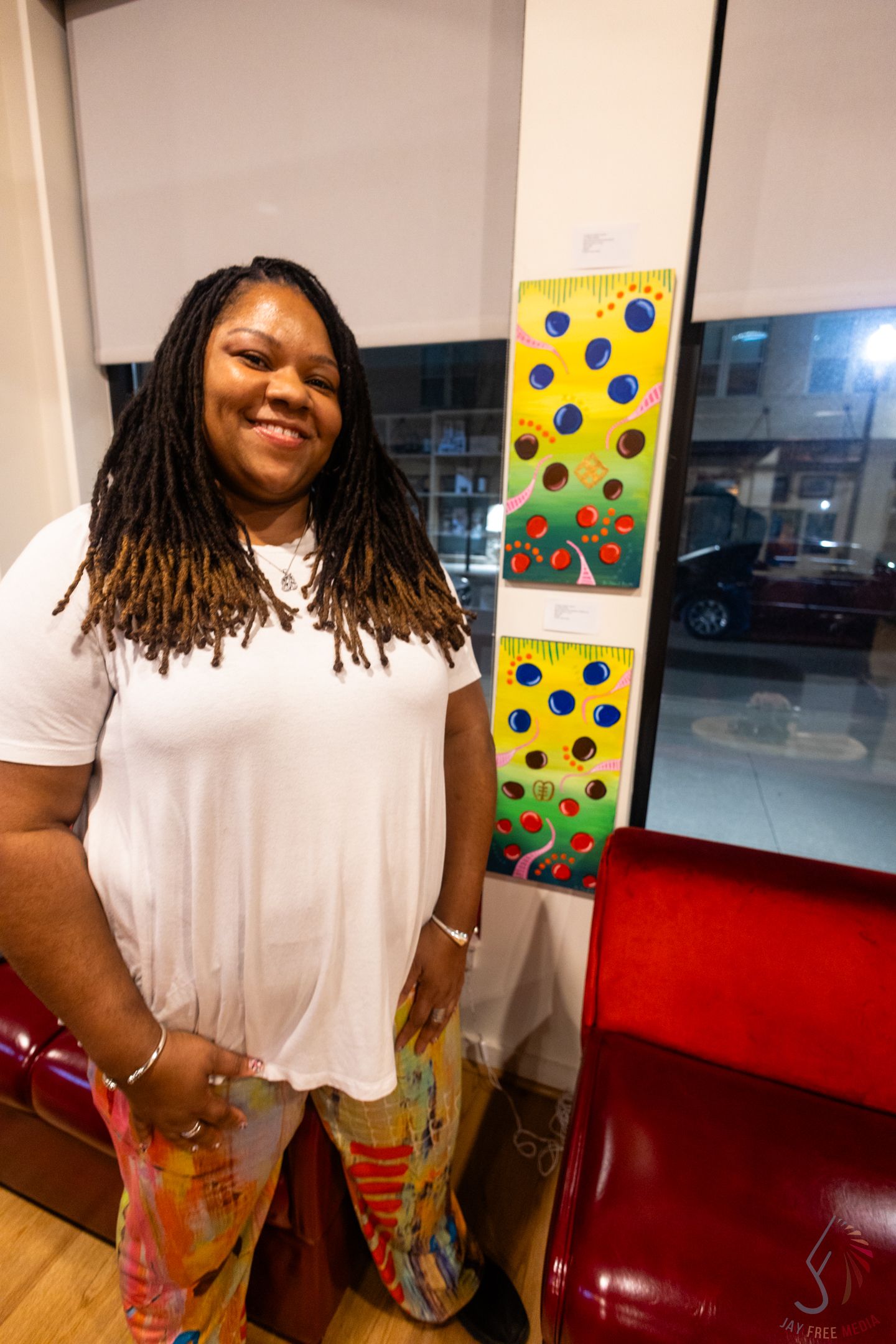 Smiling woman with locs in white tee and colorful pants stands beside vibrant dotted artwork in a gallery.