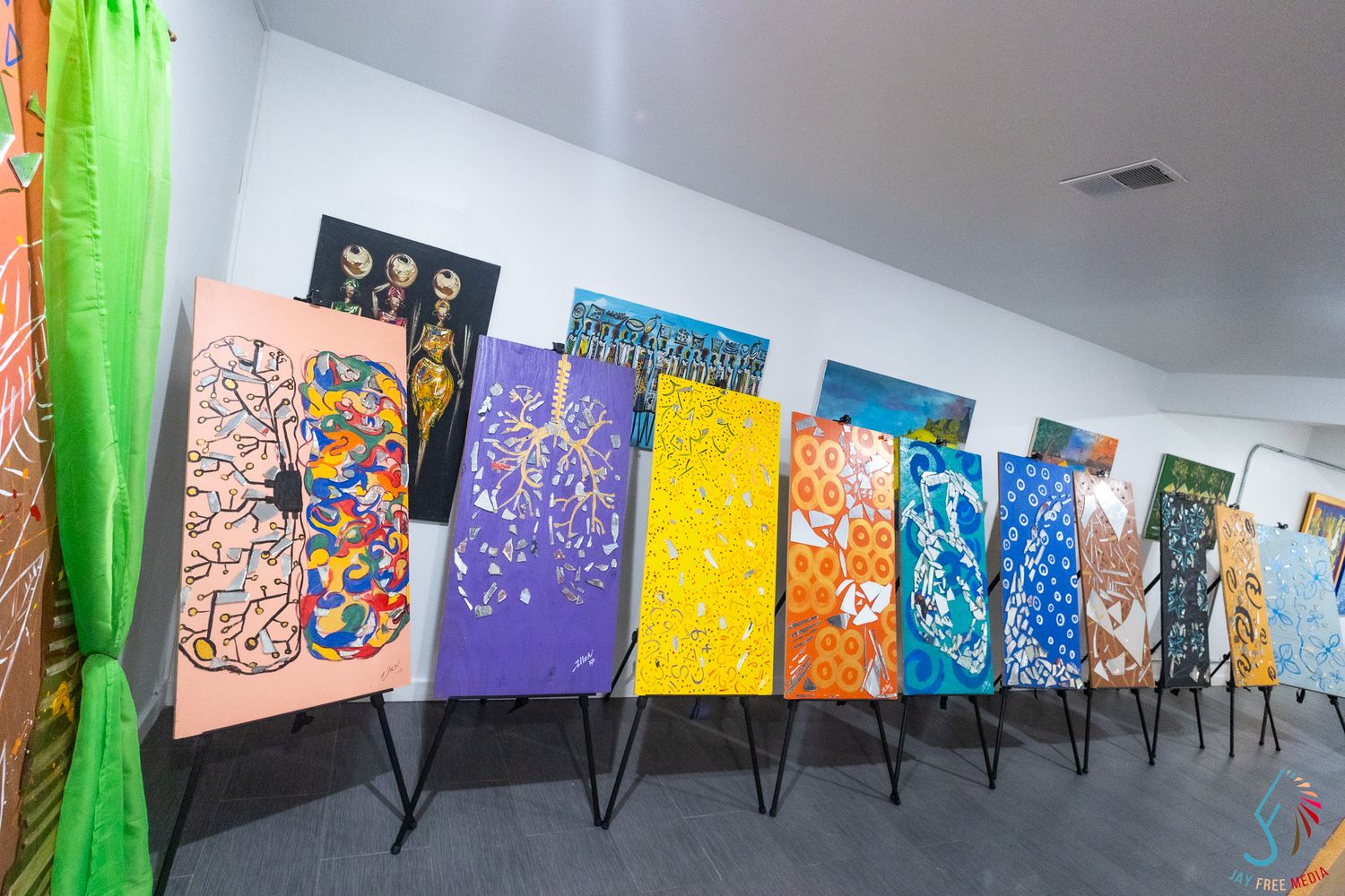 Colorful artwork paintings displayed on easels in a row inside a bright white gallery space.
