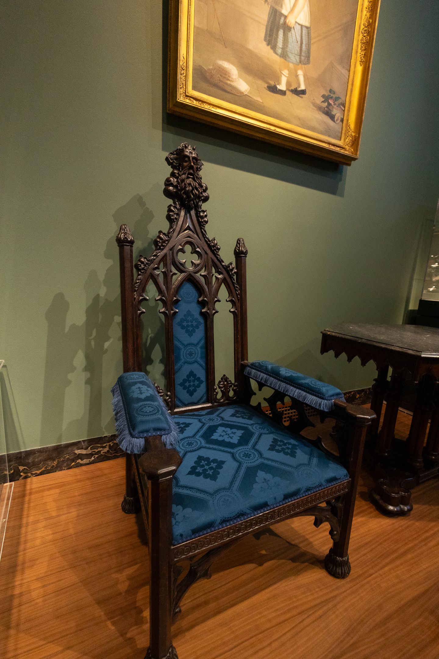 Ornate Gothic Revival wooden armchair with blue velvet upholstery displayed in a museum gallery setting.