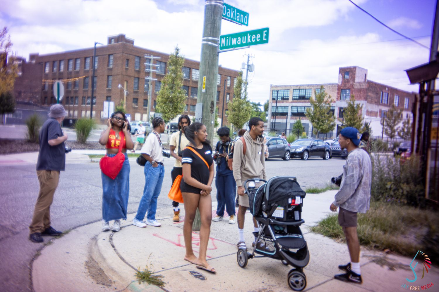 A diverse group of people gather on a urban street corner near Milwaukee Ave on a sunny day with a stroller nearby.
