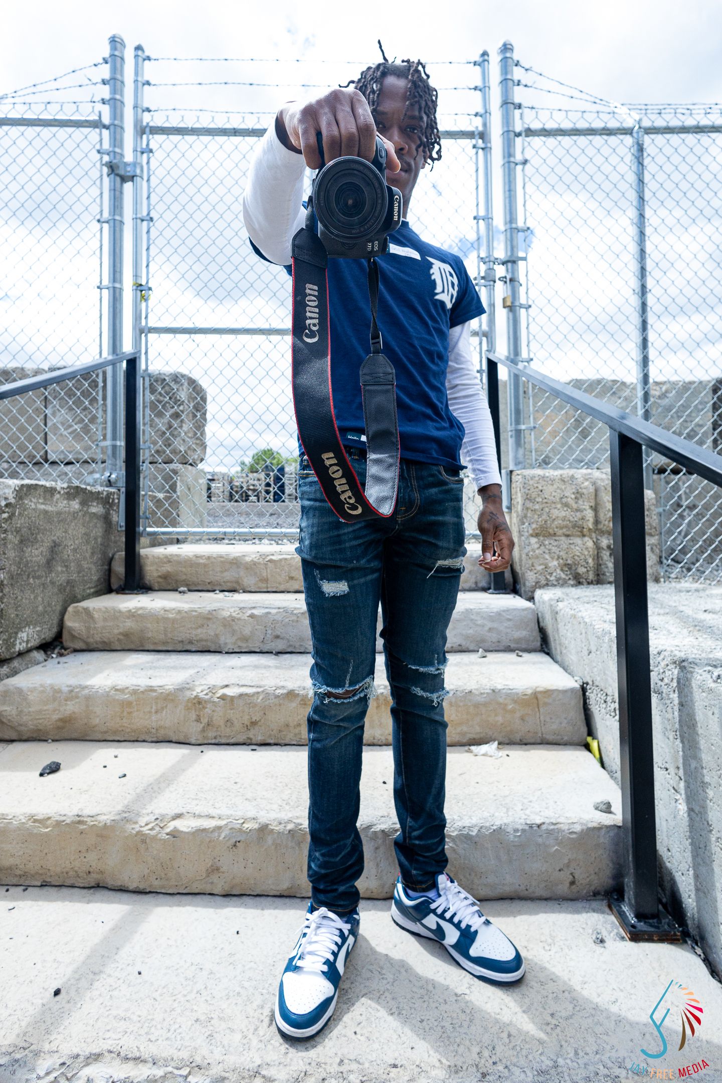 Young man in blue Detroit Tigers jersey pointing Canon camera at viewer on outdoor concrete steps.