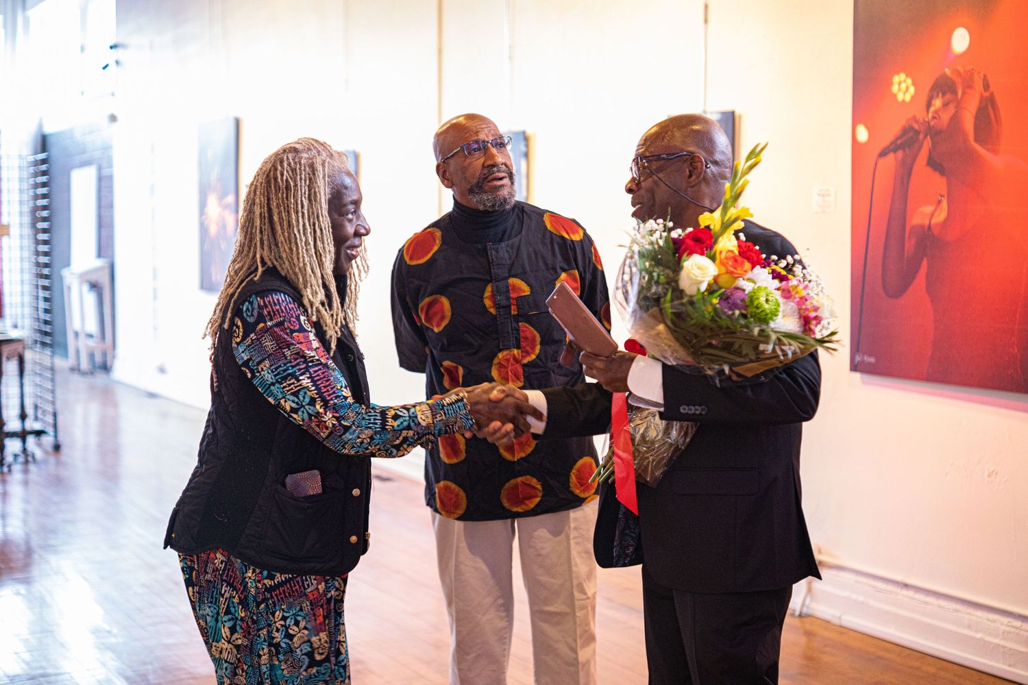 Three people greet each other warmly at an art gallery opening, one holding colorful flowers.