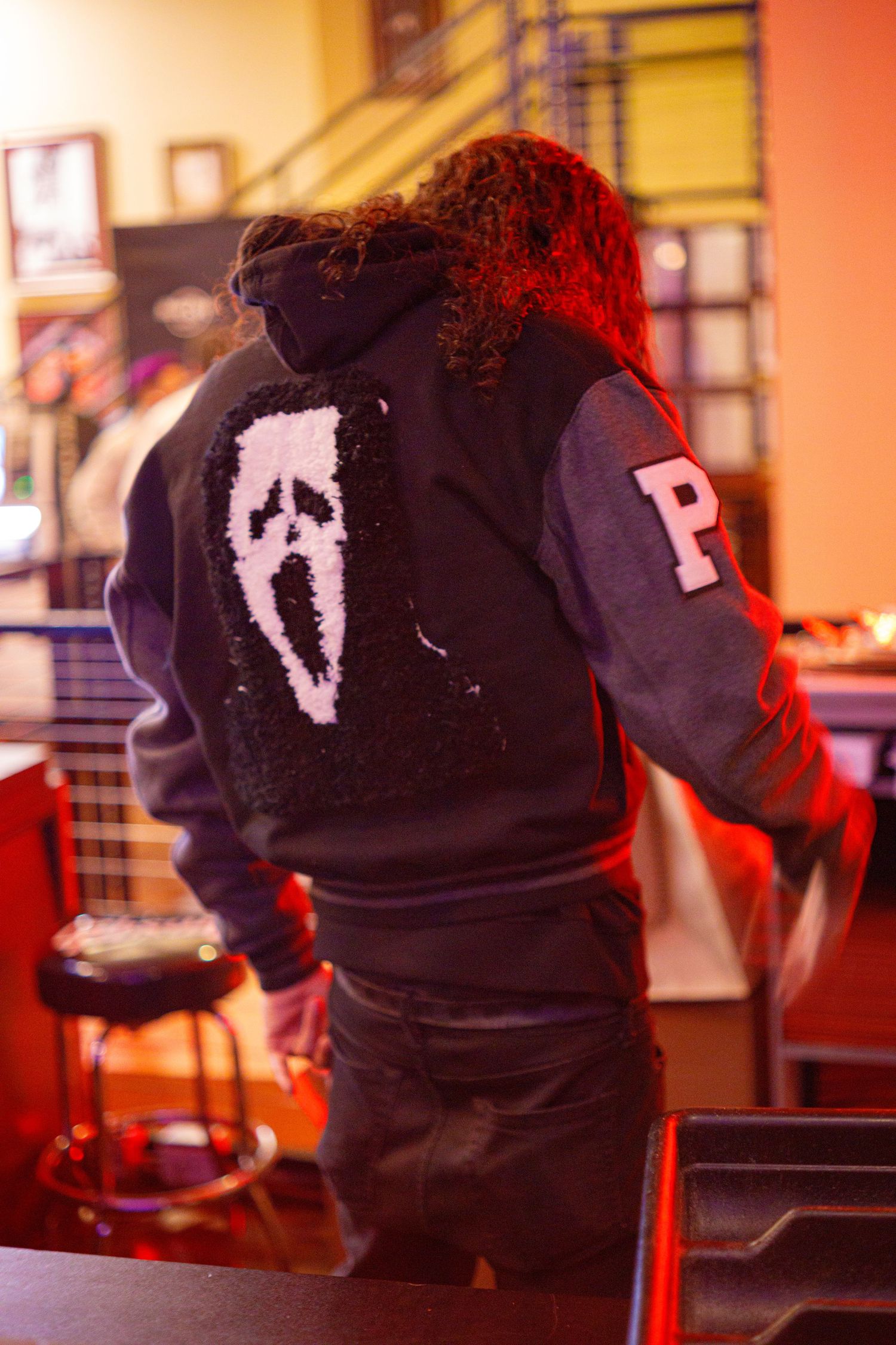 Person wearing a black hoodie with Ghostface design from Scream and letter P patch, lit by red lighting.