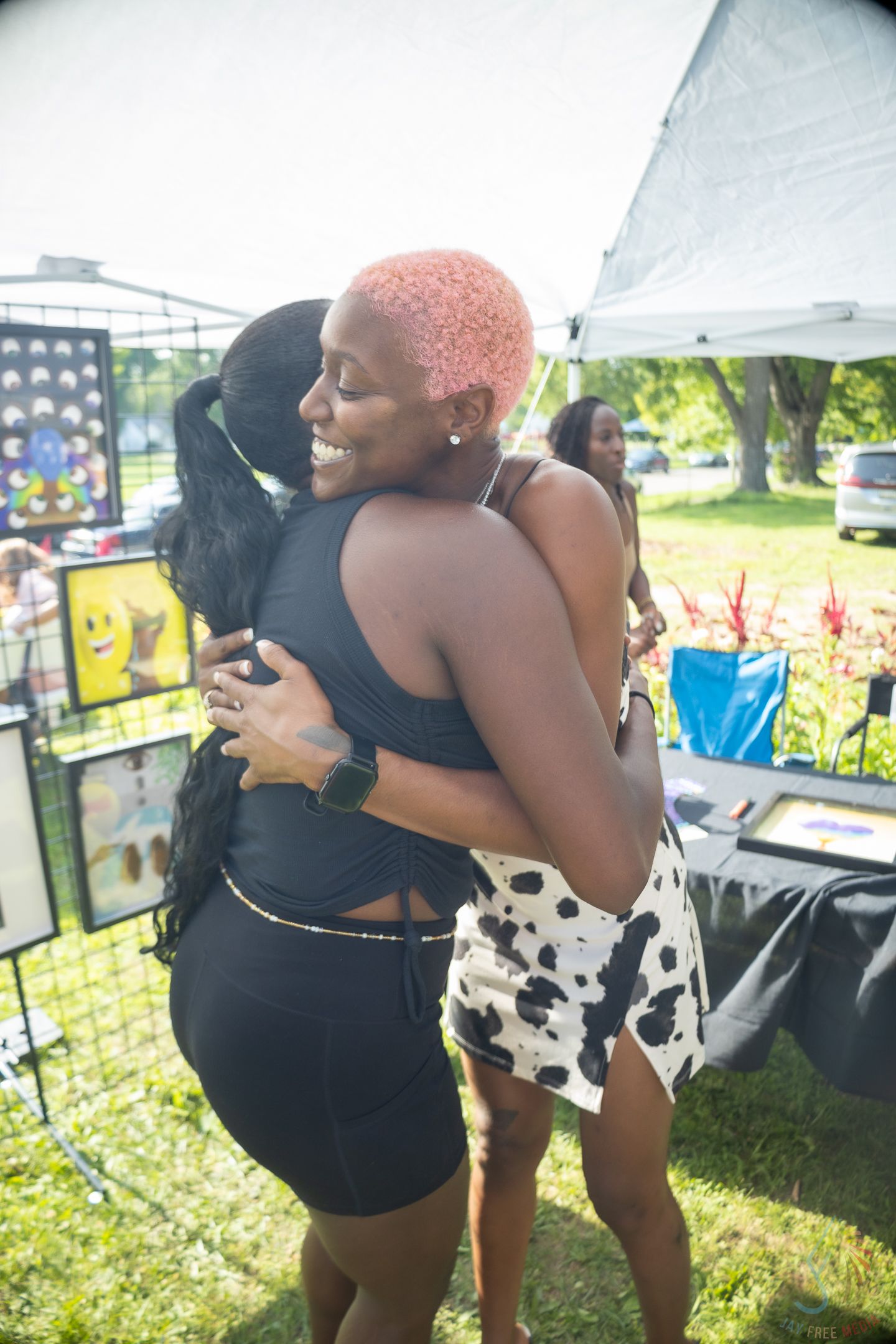 Two women share a warm hug at an outdoor art fair, one with pink hair wearing a cow-print skirt.