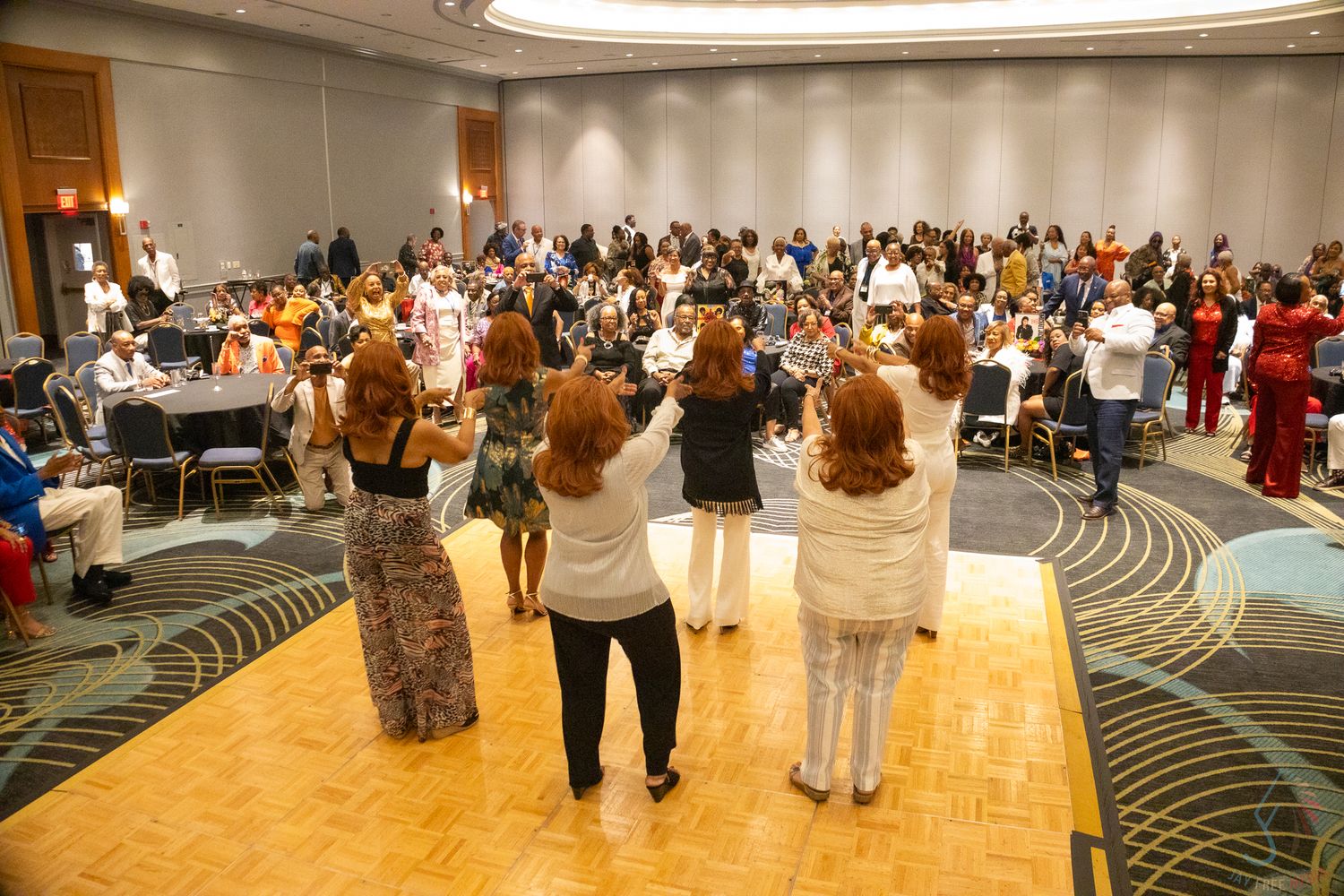 Women standing before a large crowd in a hotel ballroom during a conference or celebratory event.