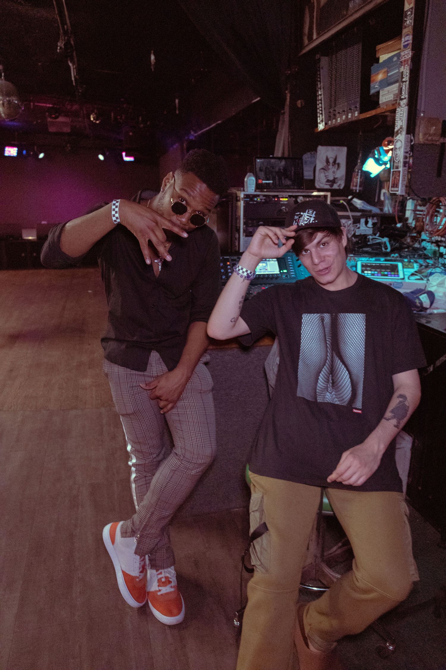 Two young men posing in a dimly lit music venue, wearing streetwear and snapback caps.