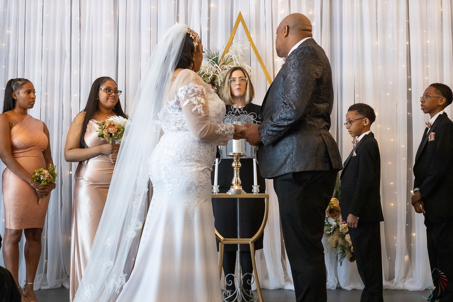 A bride and groom exchange vows at an elegant wedding ceremony with a gold geometric arch backdrop and wedding party.
