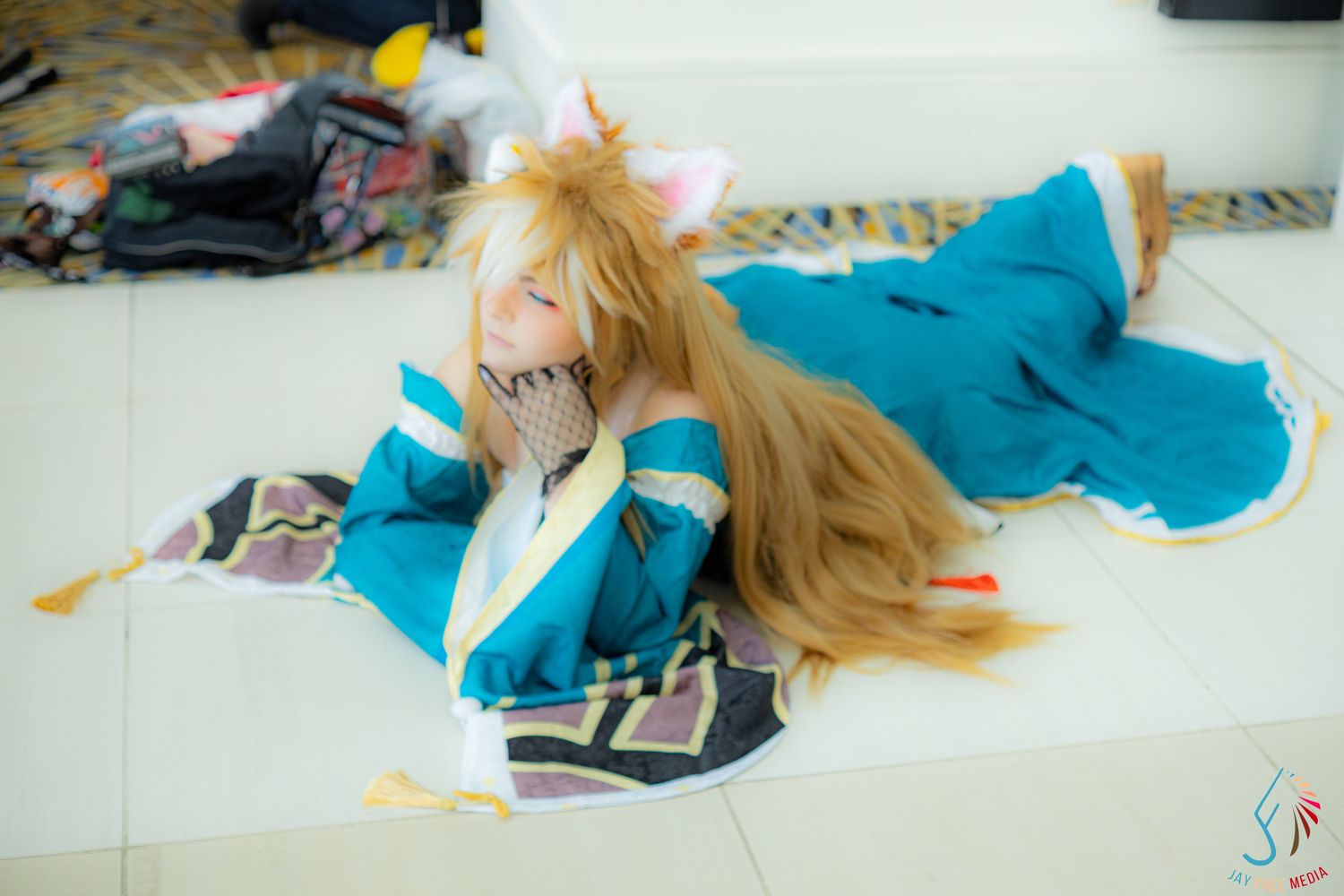 Cosplayer in a blue anime costume with long blonde wig posing on the floor at a convention.