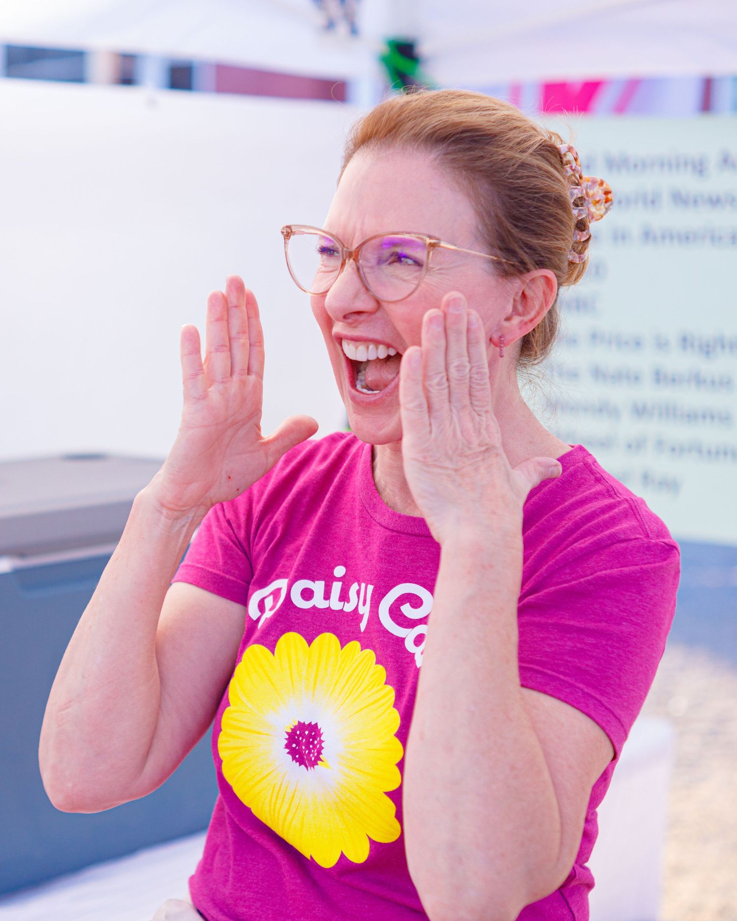 Excited redhead woman in a pink Daisy Co t-shirt laughing at an outdoor event.