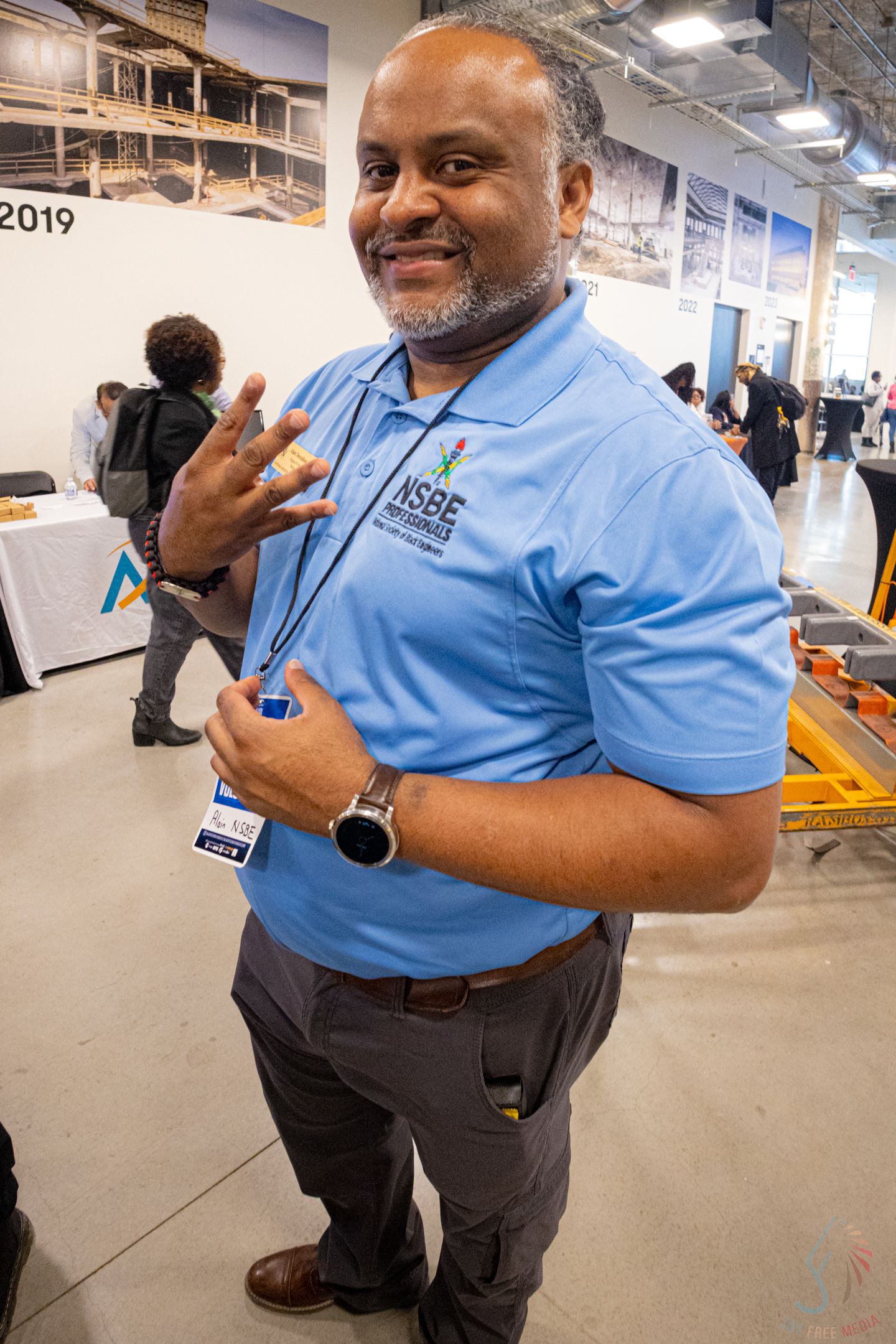 Man in blue NSBE polo shirt smiling and gesturing at a 2019 conference event.
