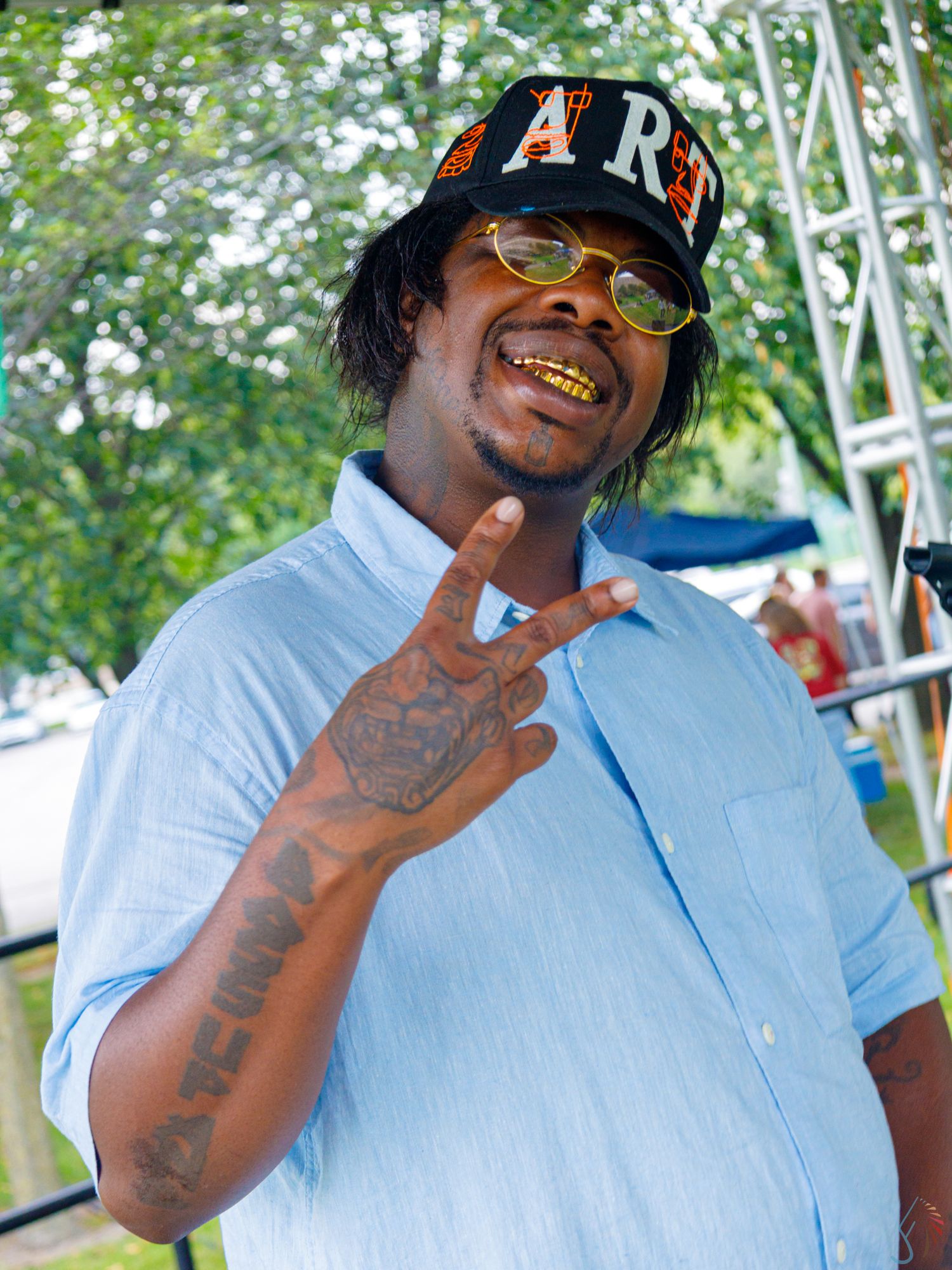 Man with gold teeth, tattoos, yellow sunglasses, and a black cap flashing a peace sign outdoors.