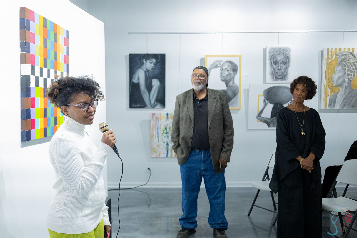 Three people stand in an art gallery with portraits and colorful artwork displayed on white walls behind them.