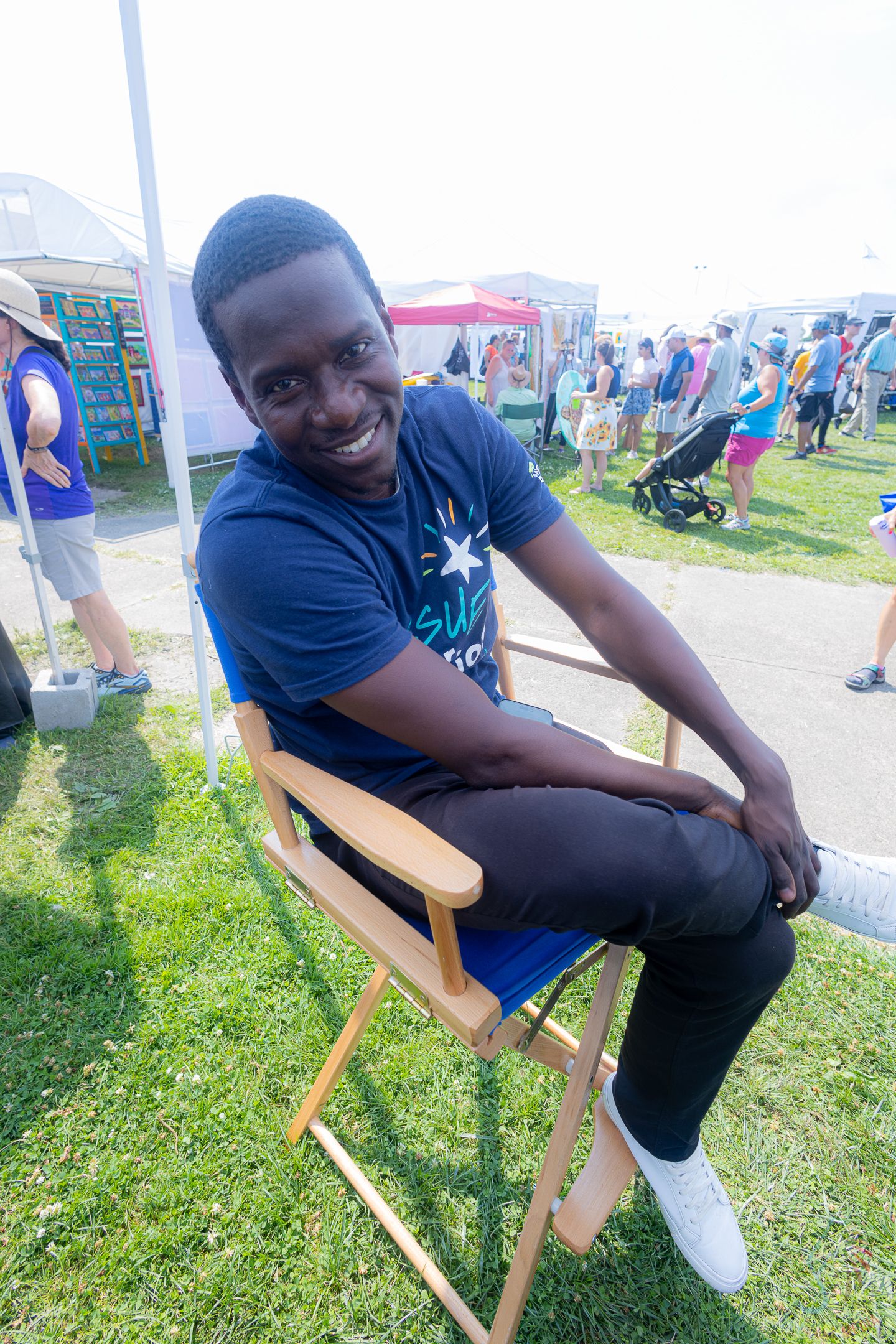 A young man in a blue t-shirt sits smiling in a folding chair at an outdoor festival.