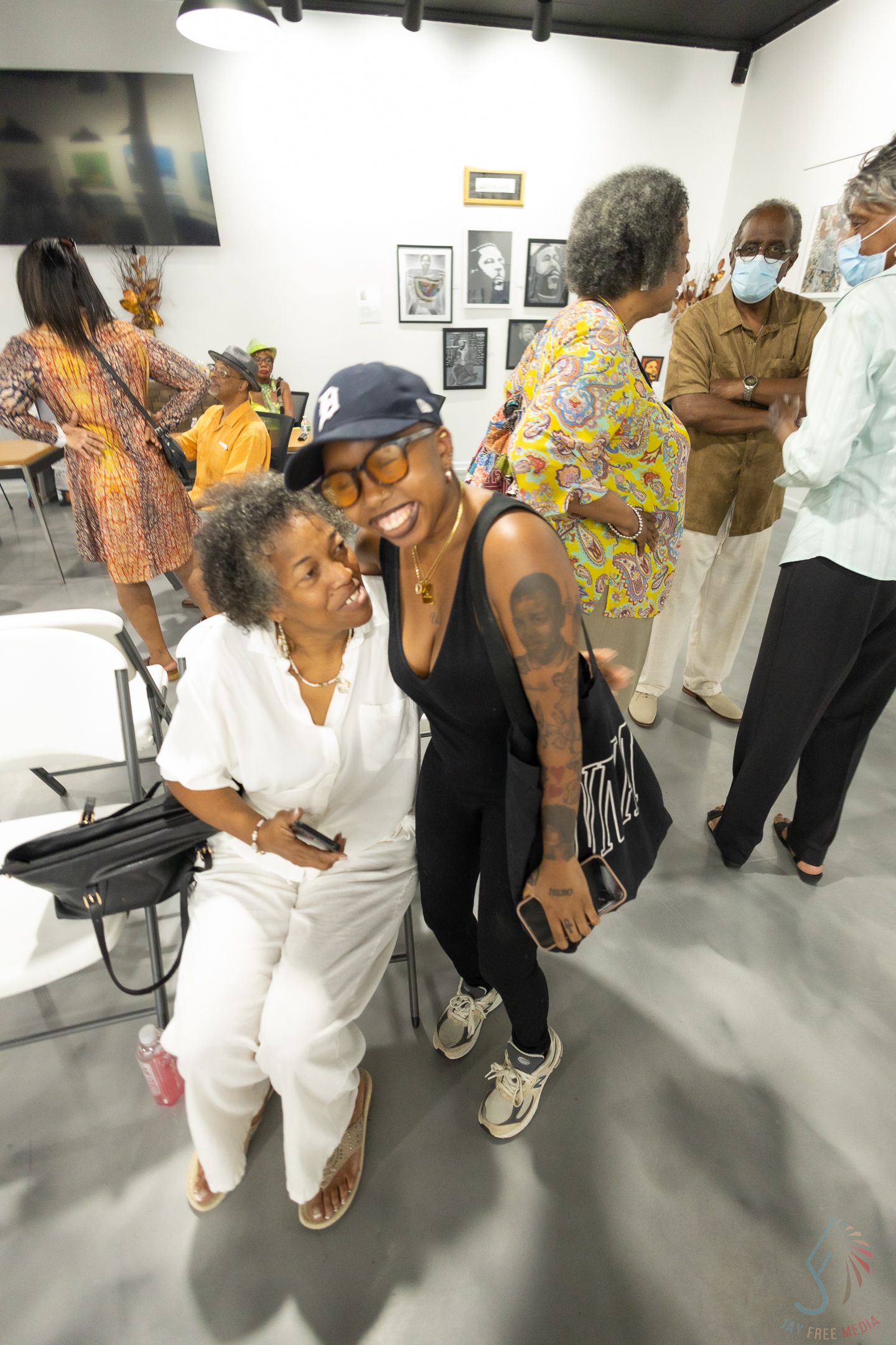 Two women smile and pose together at an art gallery event with other guests browsing artwork in the background.